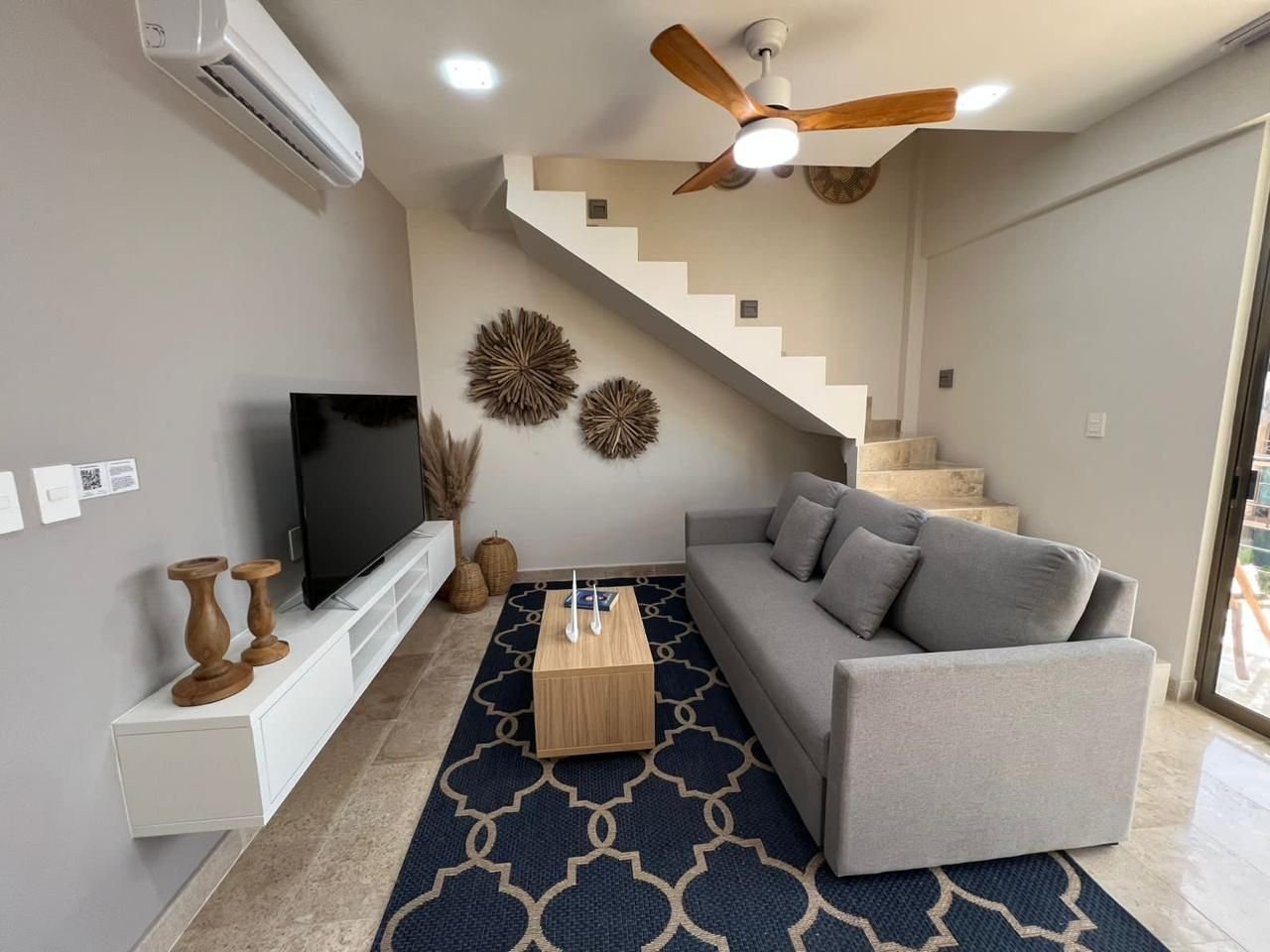 Sala con sofá gris, televisión, aire acondicionado y escalera